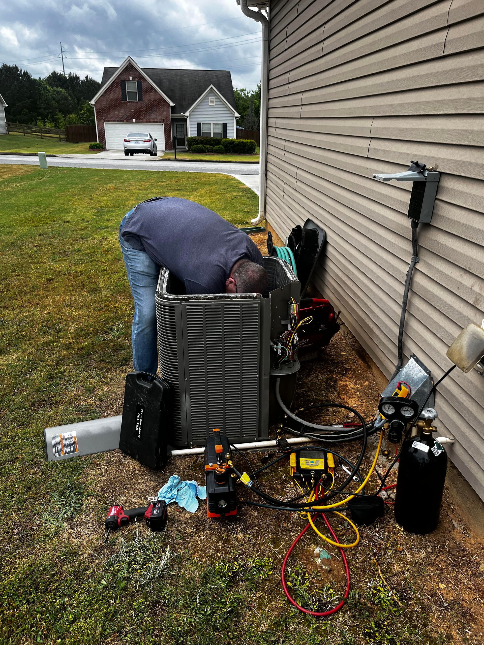 Man in blue shirt inspecting outdoor air conditioner beside a brick house