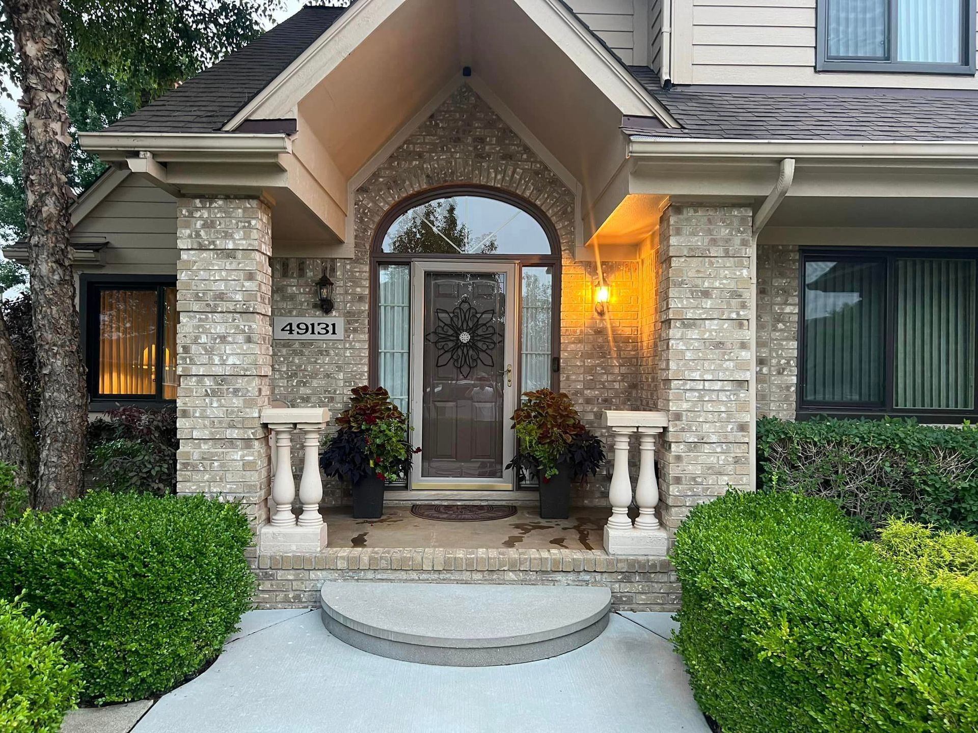 Front entrance of a stone house with arched doorway, lit porch, and neatly trimmed green hedges
