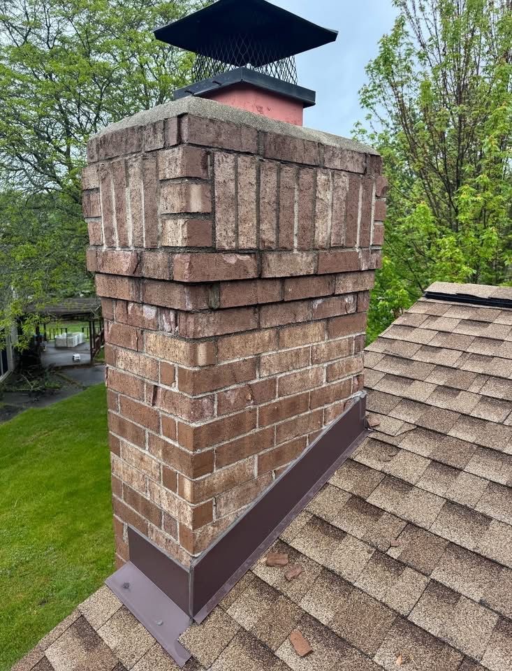 Brick chimney on a shingle roof, with a metal cap and flashing. Green trees in the background.