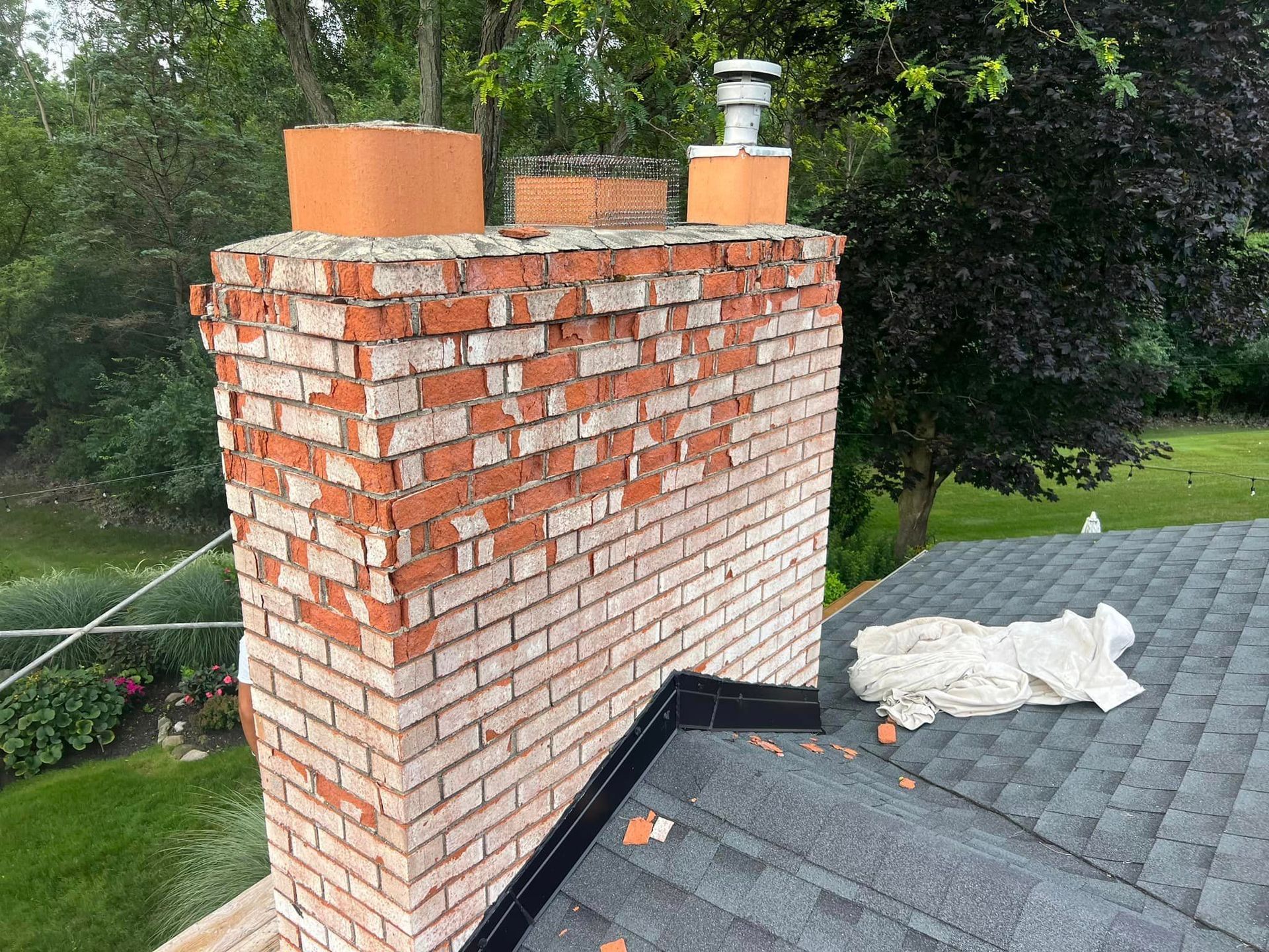 Damaged brick chimney with weathered bricks. Crumbling mortar. Located on a roof, with green foliage in the background.