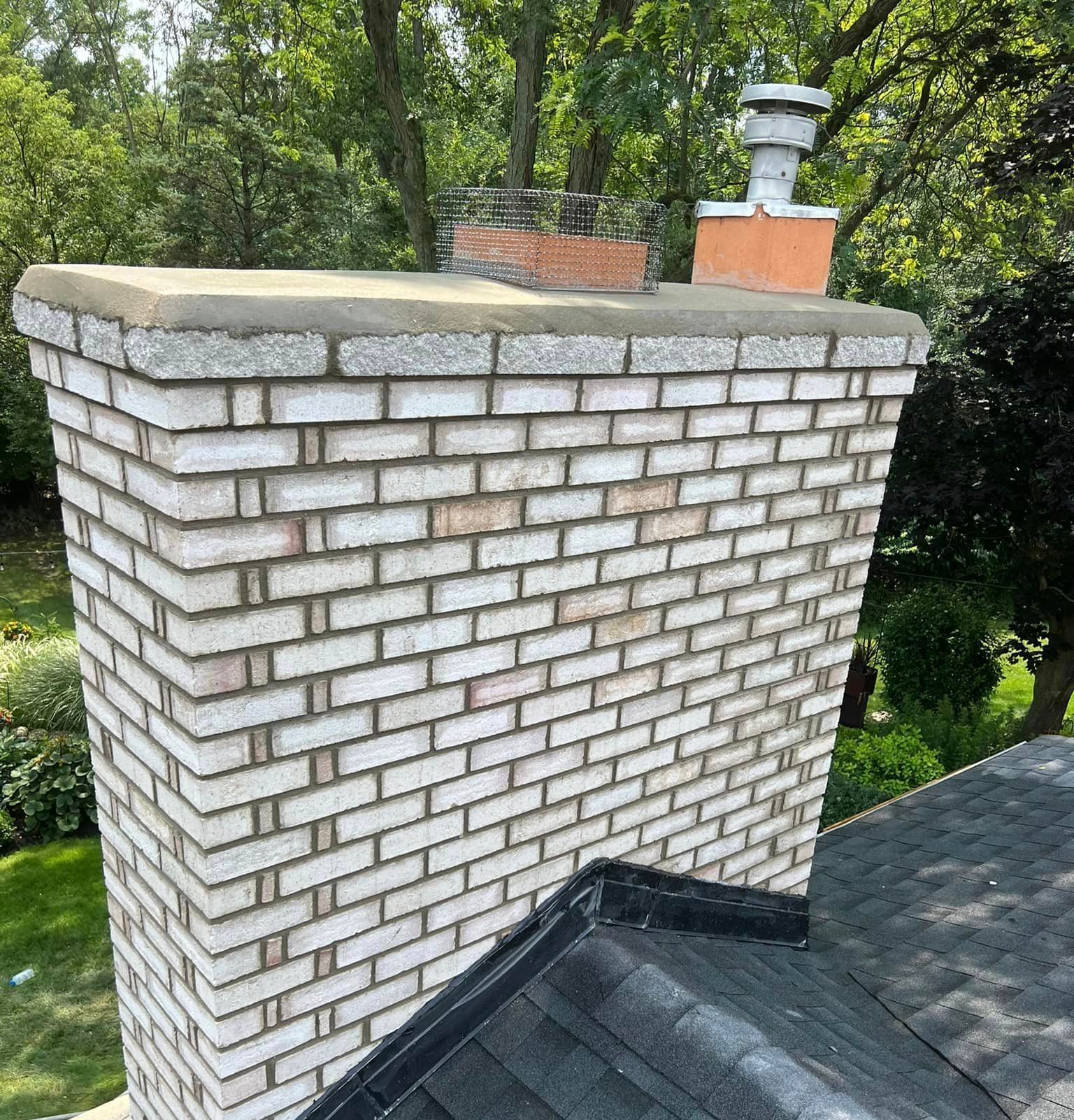 Brick chimney on a roof, with gray cap and a smaller chimney. Trees in the background.