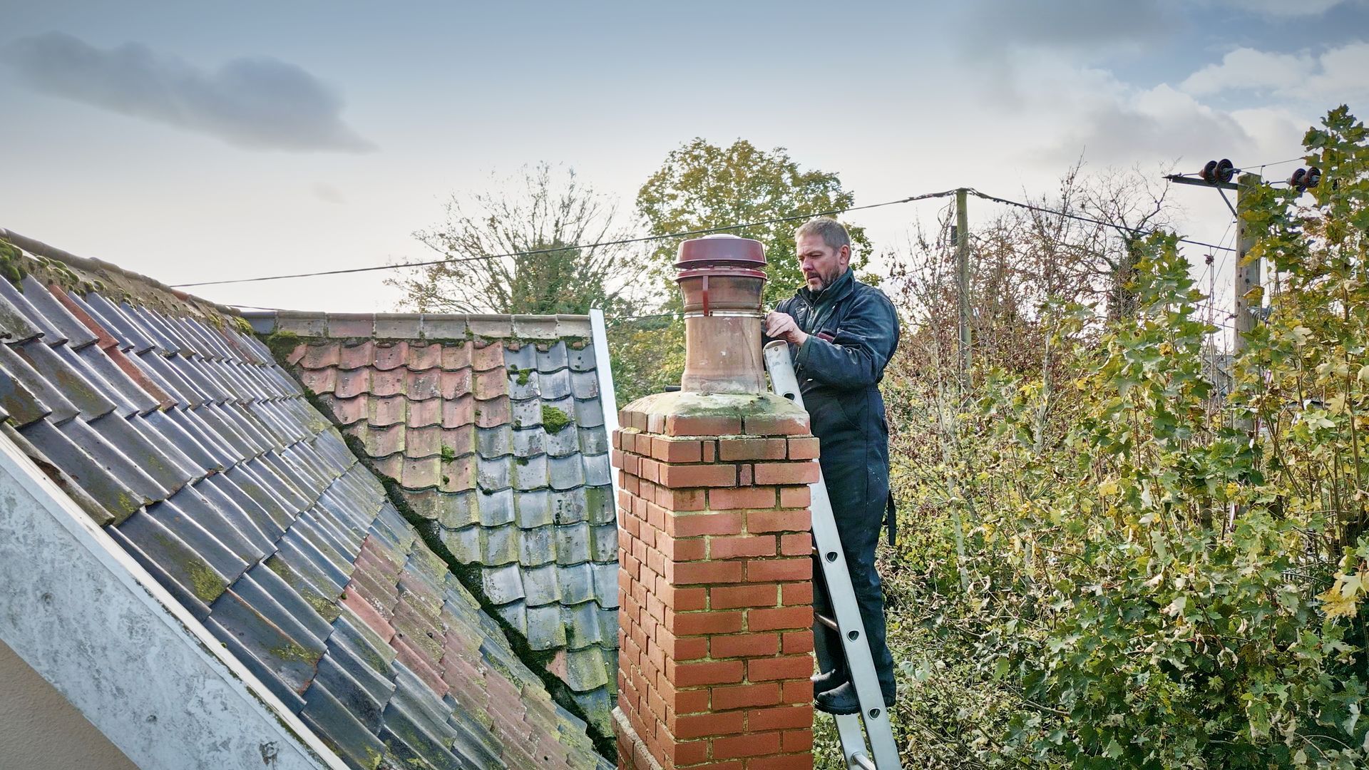 A chimney sweeps on a ladder performing a chimney repair service on a residential roof.