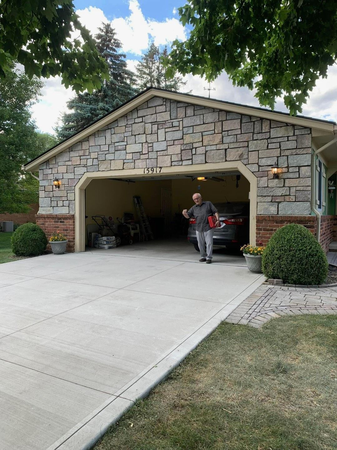 Man exits a garage with stone facade. A car is parked inside; green shrubbery and a concrete driveway are in view.