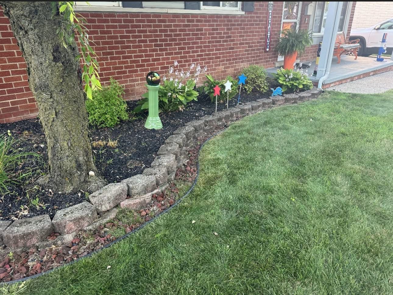 Brick house garden bed with black mulch, colorful flower stakes, and a green bird feeder beside a lawn