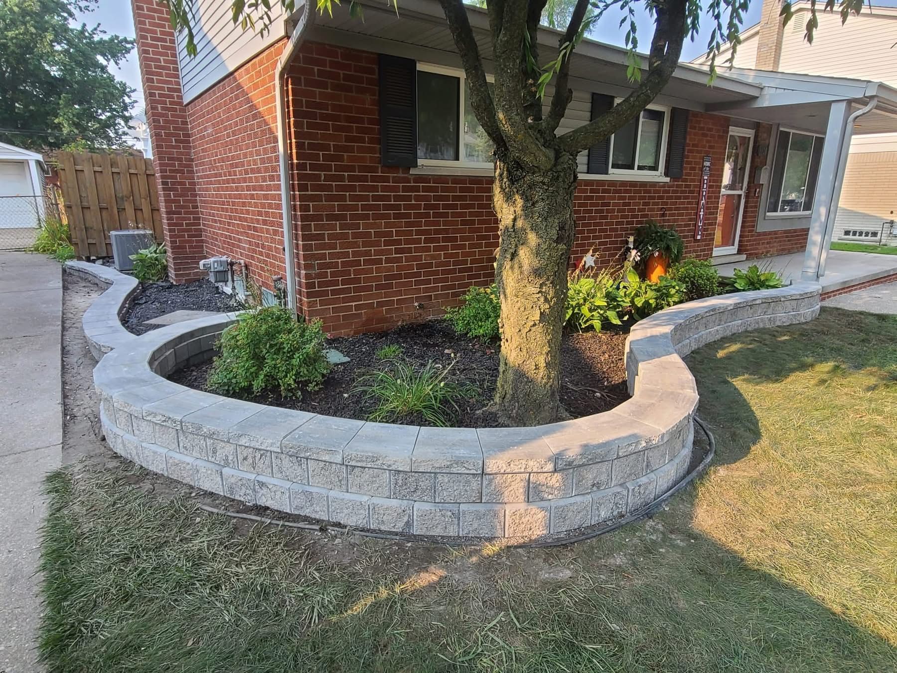 Curved stone retaining wall around a brick house tree bed with mulch and plants