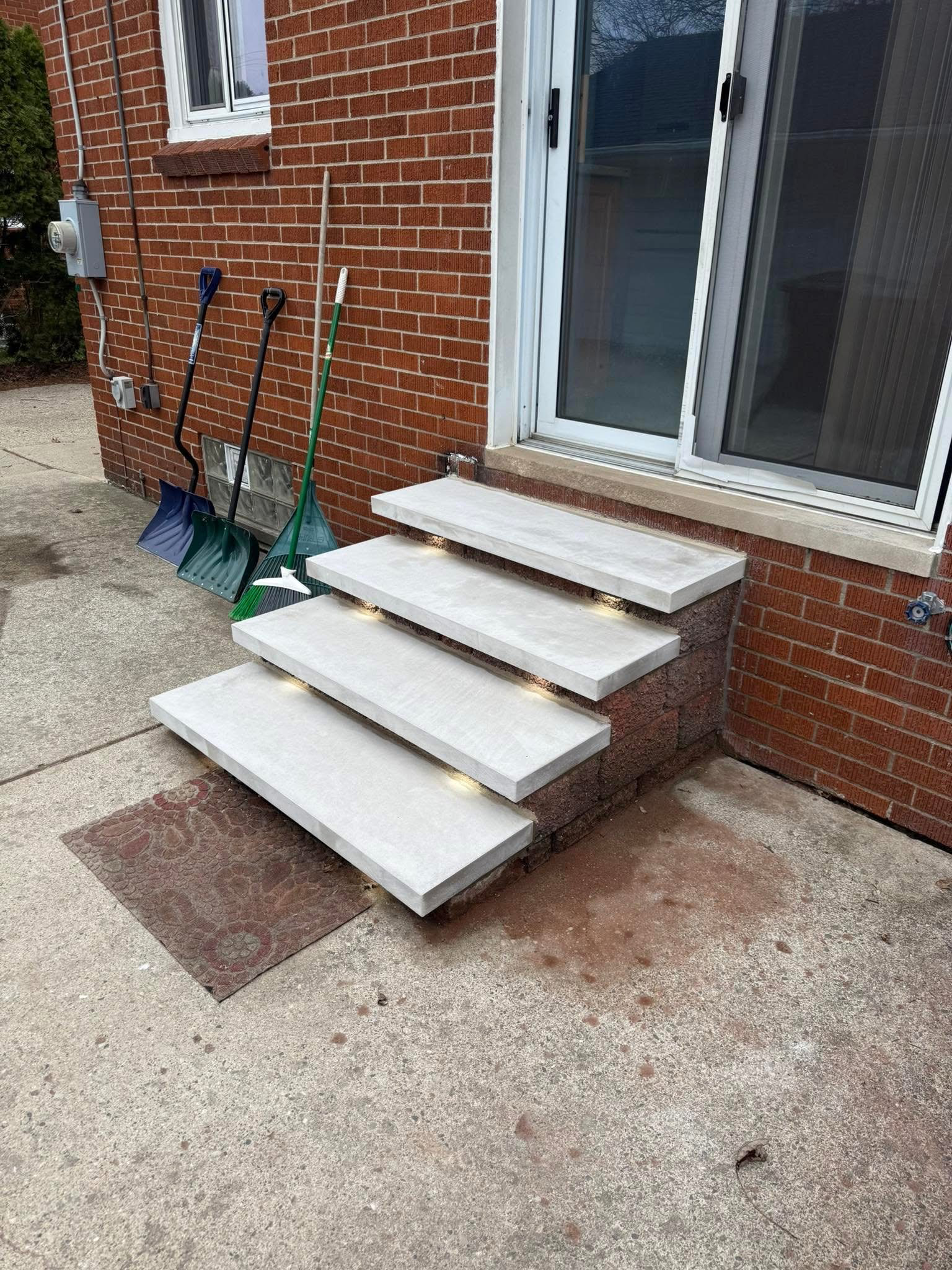 White portable steps leading to a brick house door, with shovels leaning against the wall.