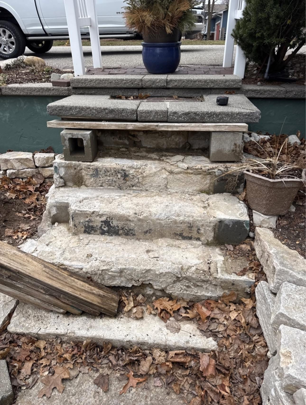 Broken stone steps leading to a raised planter beside a sidewalk, with fallen leaves and a blue pot nearby