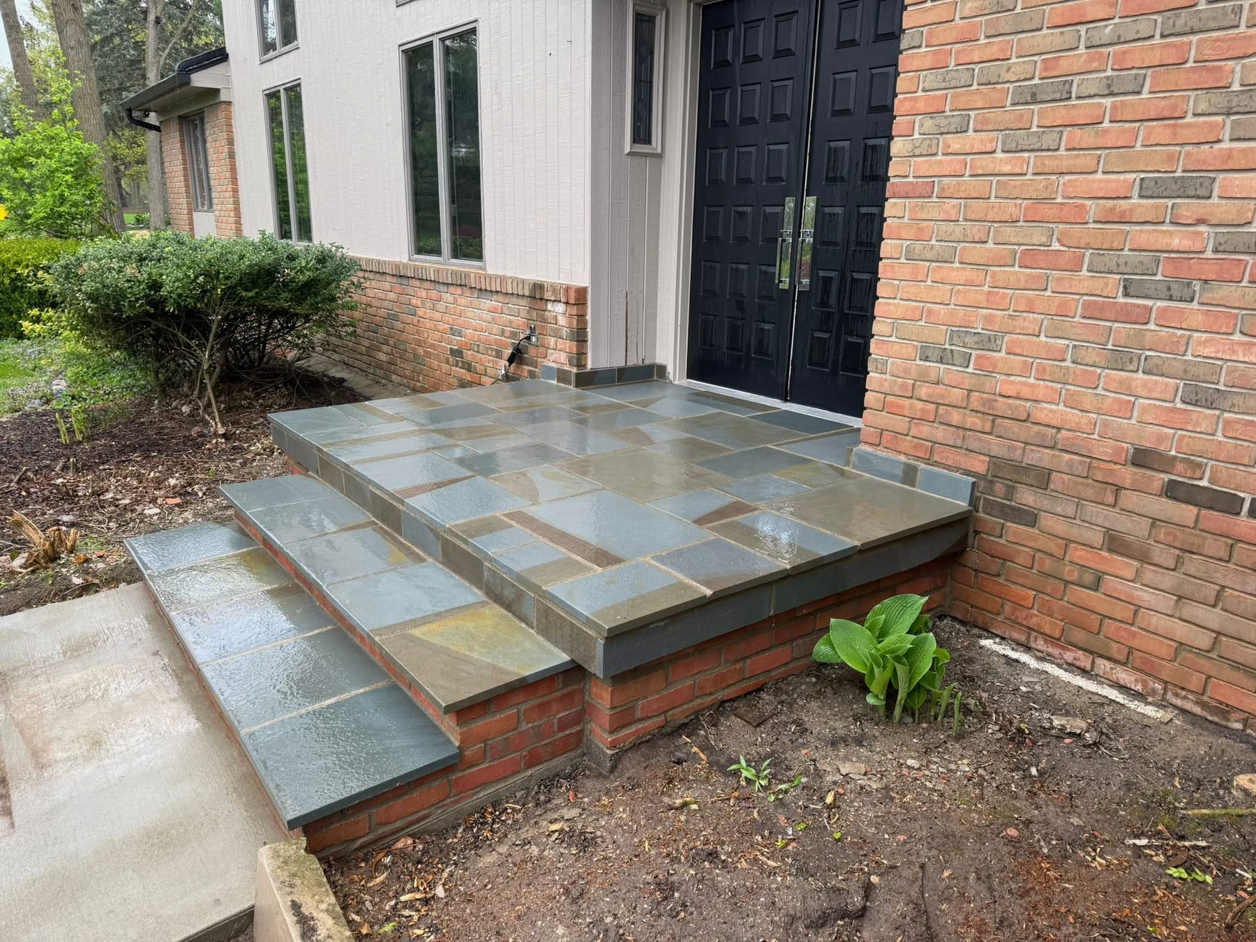 Front entrance with brick and white siding, black door, and wide slate-tiled steps with a side ramp.