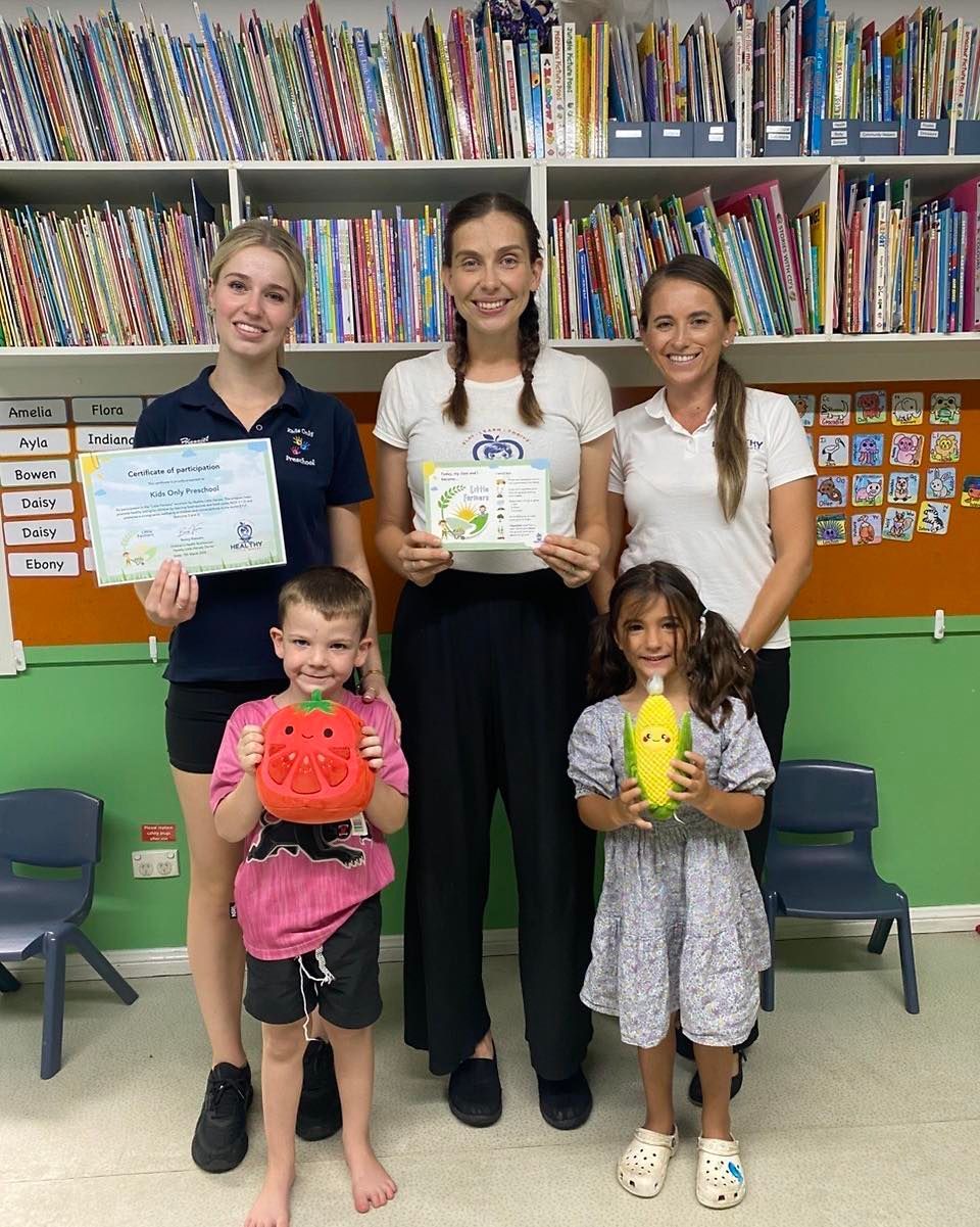 Three women and two children pose in a classroom holding certificates and colorful stuffed toys.