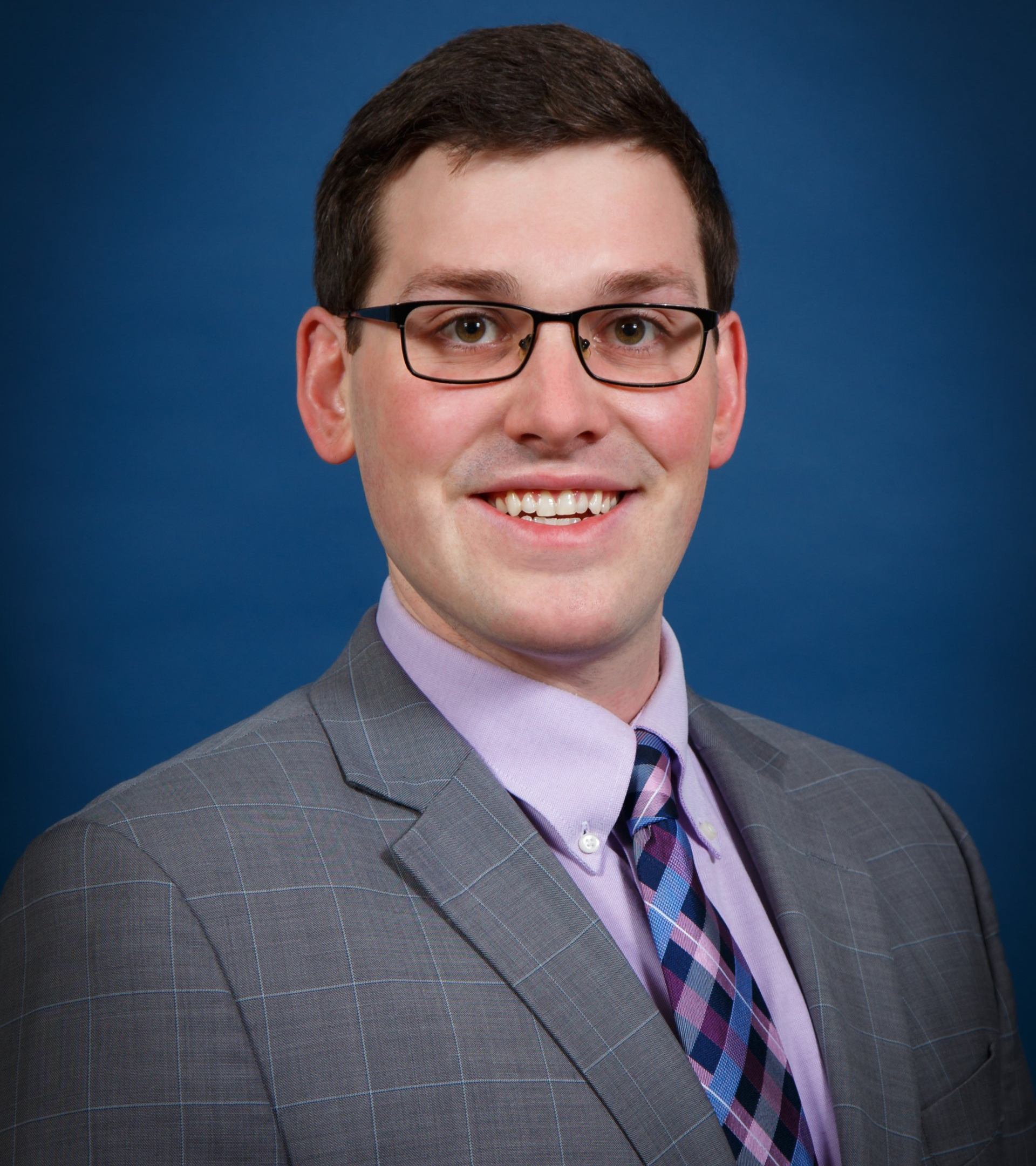 Man with glasses and suit smiling against a blue backdrop.