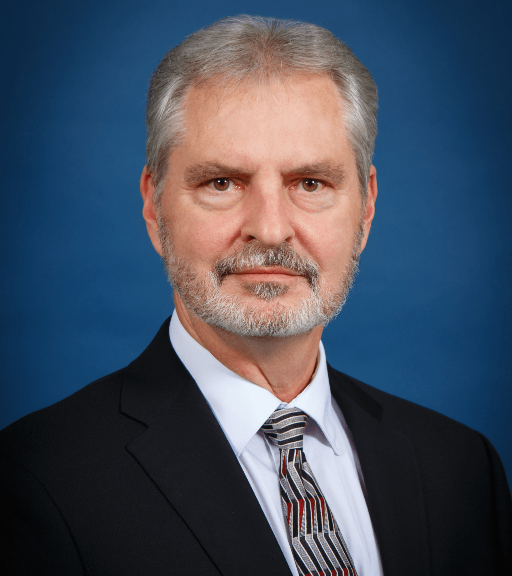 Man with gray hair and beard, wearing a suit and tie, against a blue background.