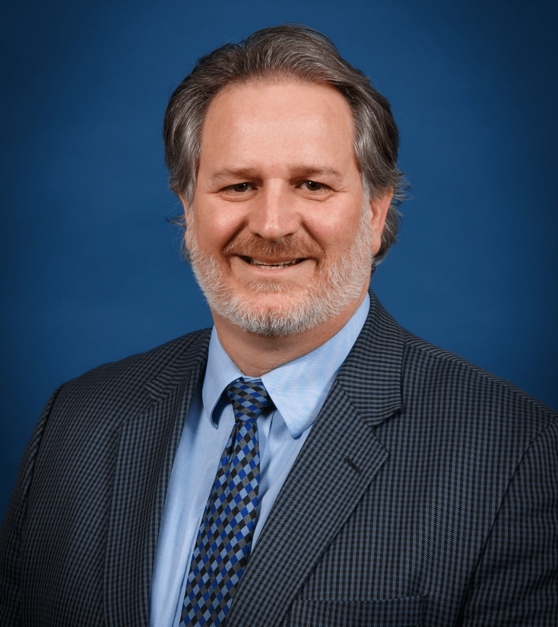 Man in suit smiling, with a grey beard, blue tie, and a blue background.