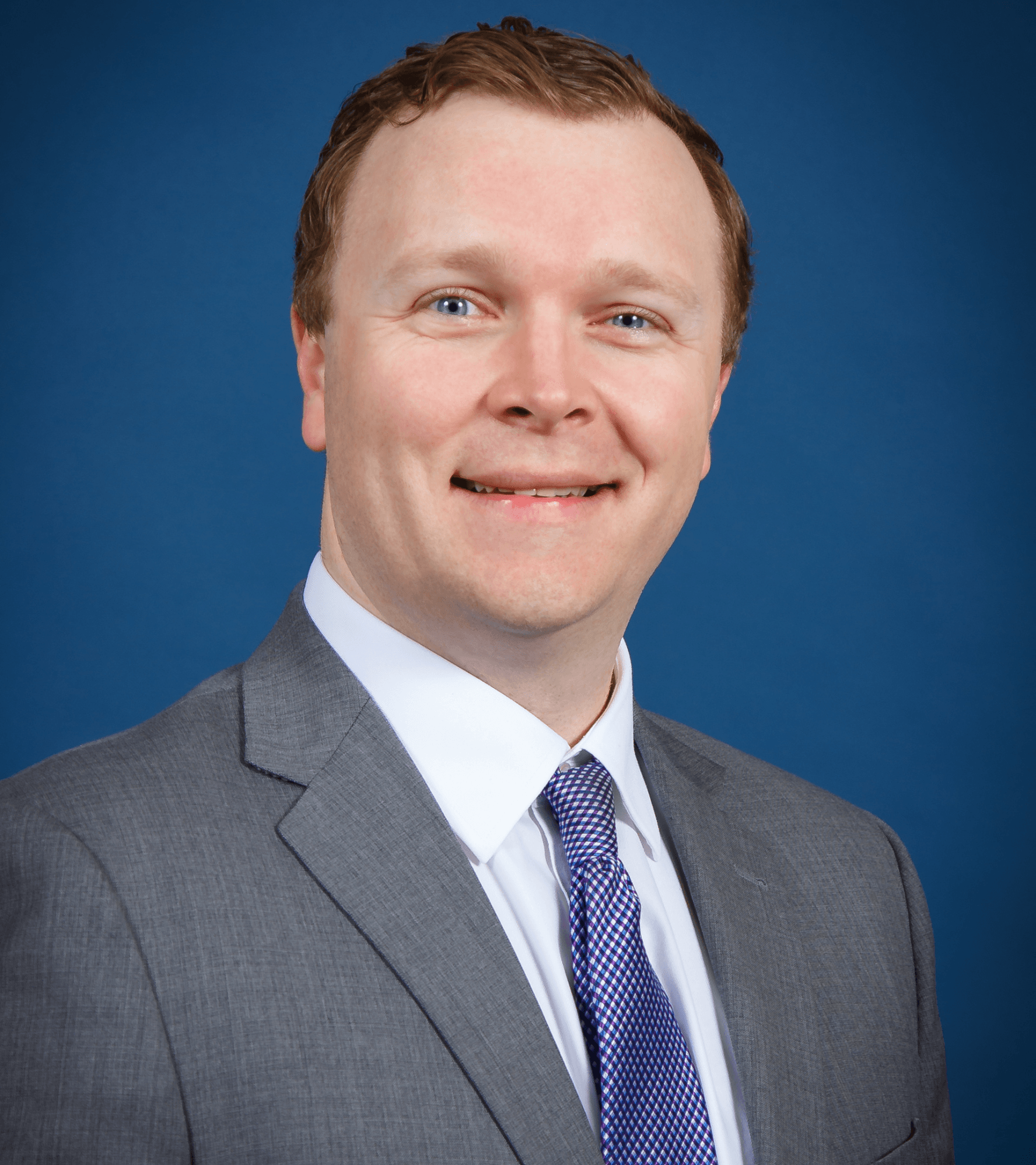 Man in gray suit and blue patterned tie smiles in front of a blue background.