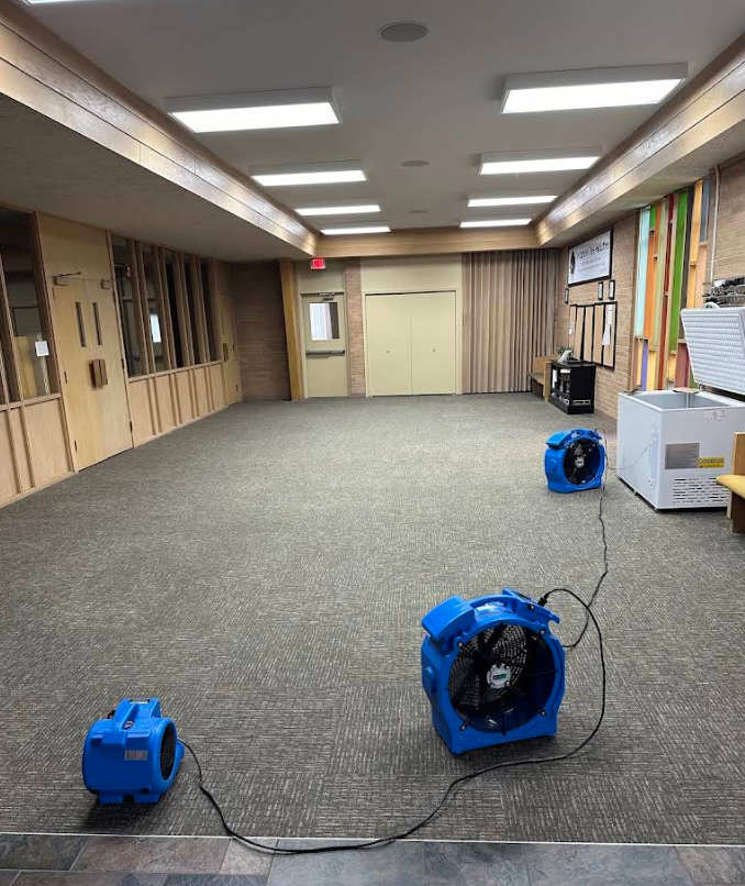 Empty carpeted room with blue fans and cords on the floor, fluorescent lights overhead.