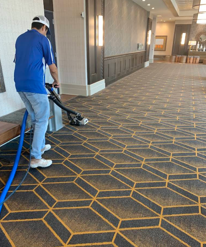 Man vacuuming a patterned hotel hallway carpet with a blue corded vacuum