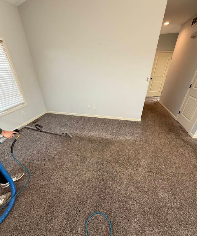 Empty beige-carpeted room with a vacuum hose at left and hallway on the right.