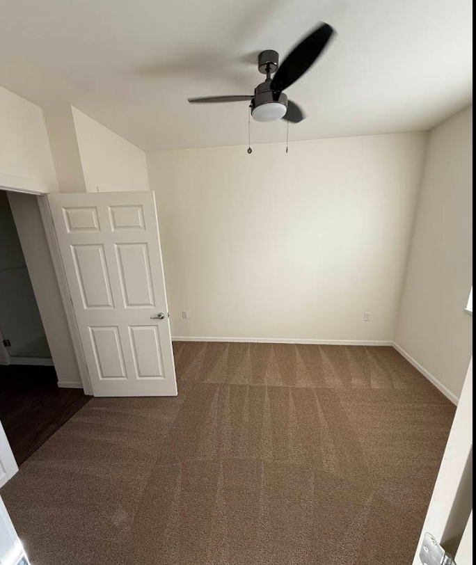 Empty beige bedroom with brown carpet, white walls, open door, and ceiling fan.