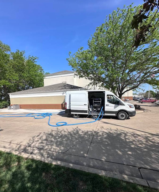 White service van with open side doors and blue hoses parked by a beige building under sunny trees