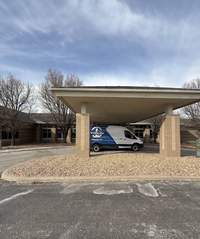White van parked under a beige covered entrance at a building, with trees and cloudy sky nearby.