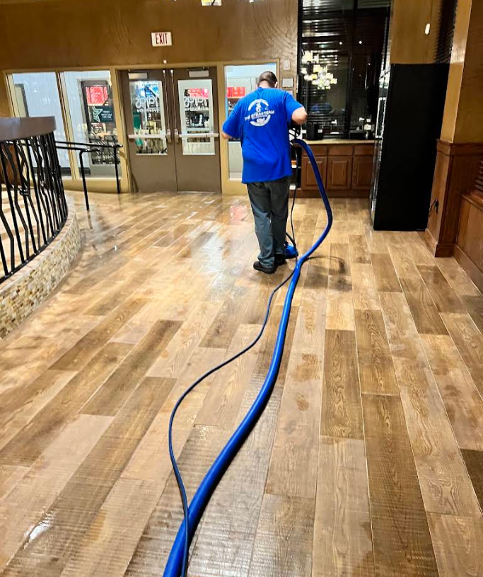 Worker in a blue shirt using a floor buffer in a wood-floored lobby, with a long blue hose trailing behind.