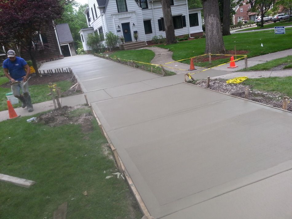 Worker smoothing wet concrete sidewalk next to a house with a green lawn.
