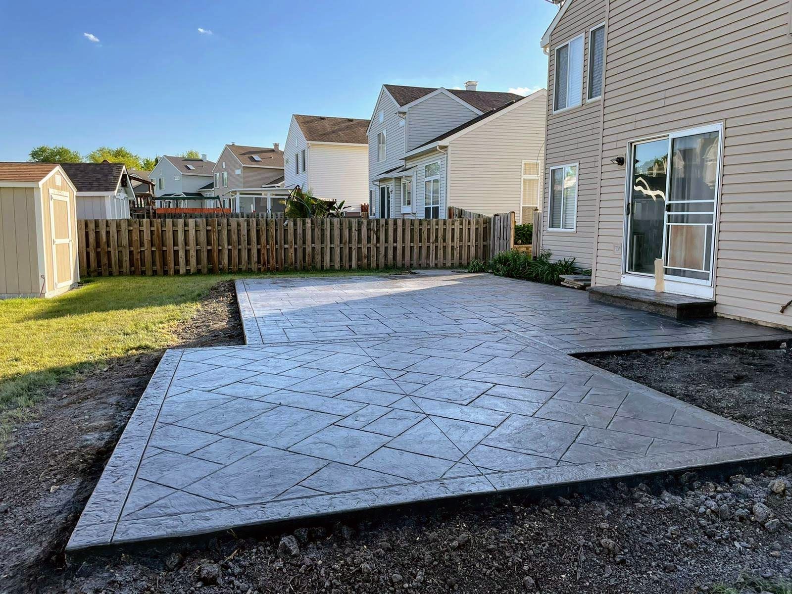 Stamped concrete patio with a decorative pattern, next to a house and yard with a fence.