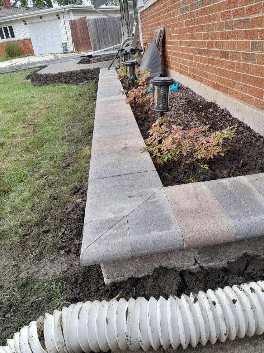 Brick retaining wall along a brick house with a flower bed and a drainage pipe in the foreground.