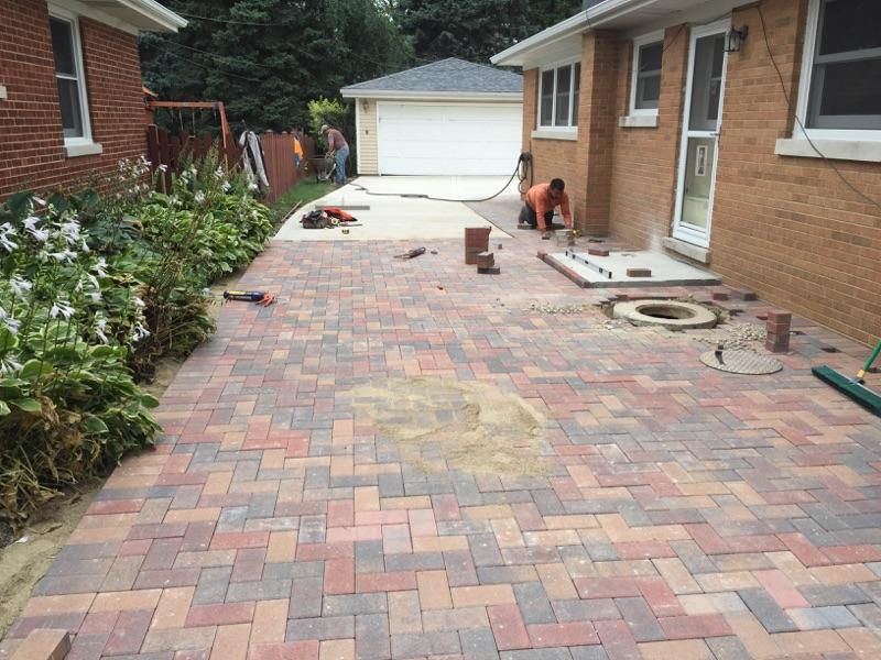 A man is working on a brick driveway in front of a brick house.
