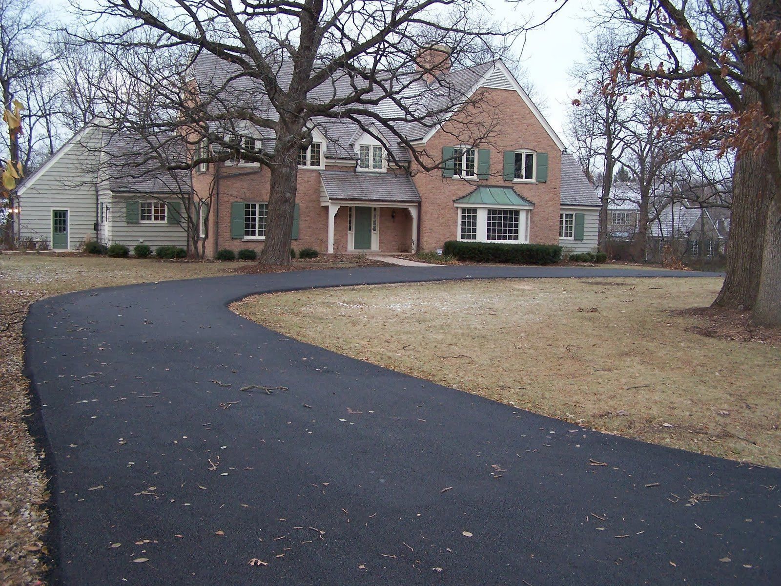 Asphalt driveway curves to a two-story brick house with green shutters and front door under a porch.