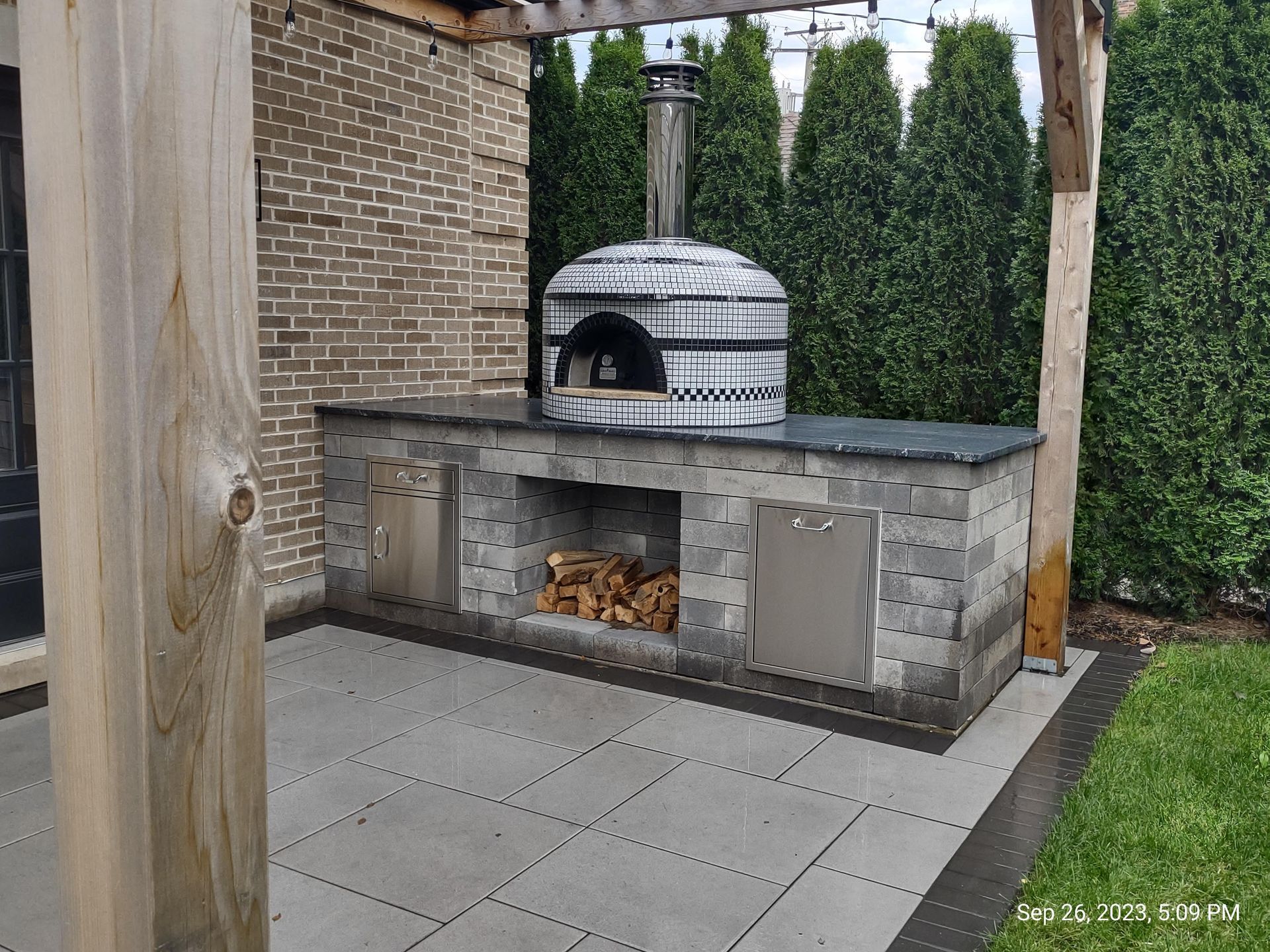 Outdoor pizza oven on a gray stone counter with a wood storage area, under a pergola, near greenery.