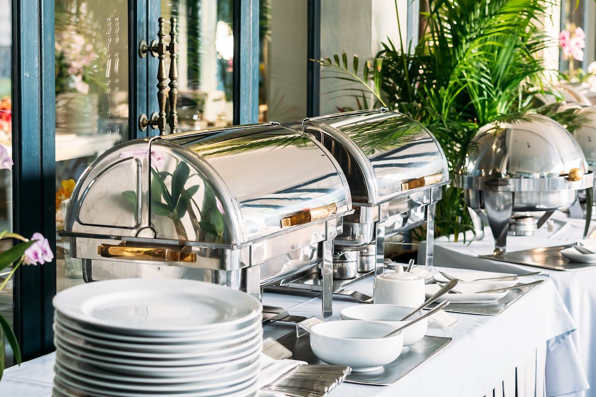 Buffet table with polished silver chafing dishes, stacked plates, and serving bowls by large windows