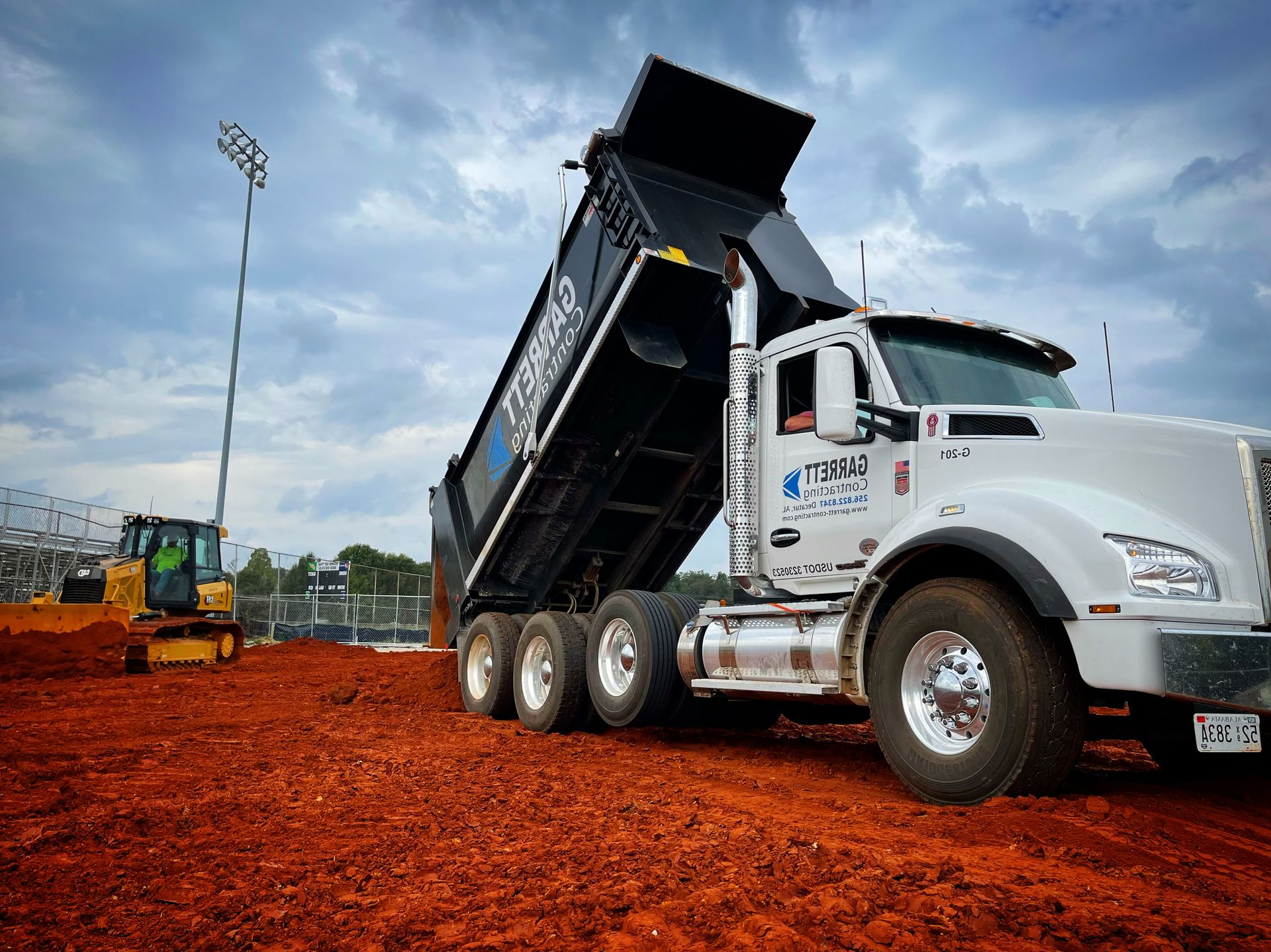 White dump truck unloading black dirt at a construction site with a bulldozer and cloudy sky