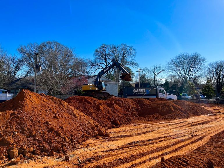 Yellow excavator, dump truck, and green loader working on a red dirt construction site.