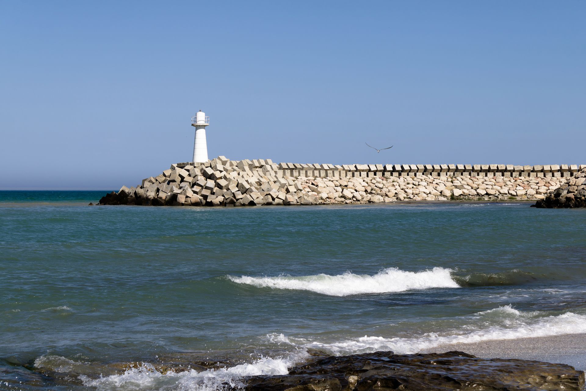 White lighthouse on rocky breakwater with sea waves crashing in foreground.