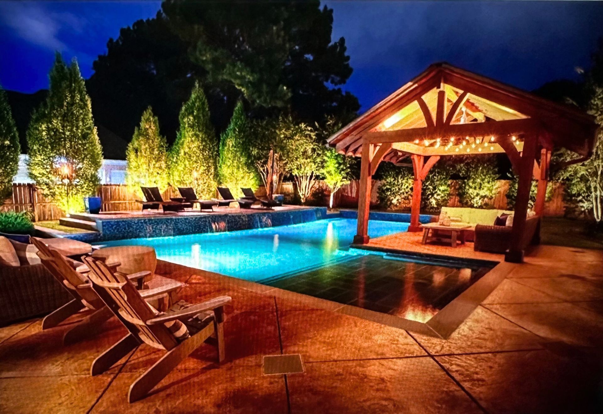 Nighttime pool scene: illuminated pool, wooden gazebo with seating, patio chairs, and landscaped with trees.