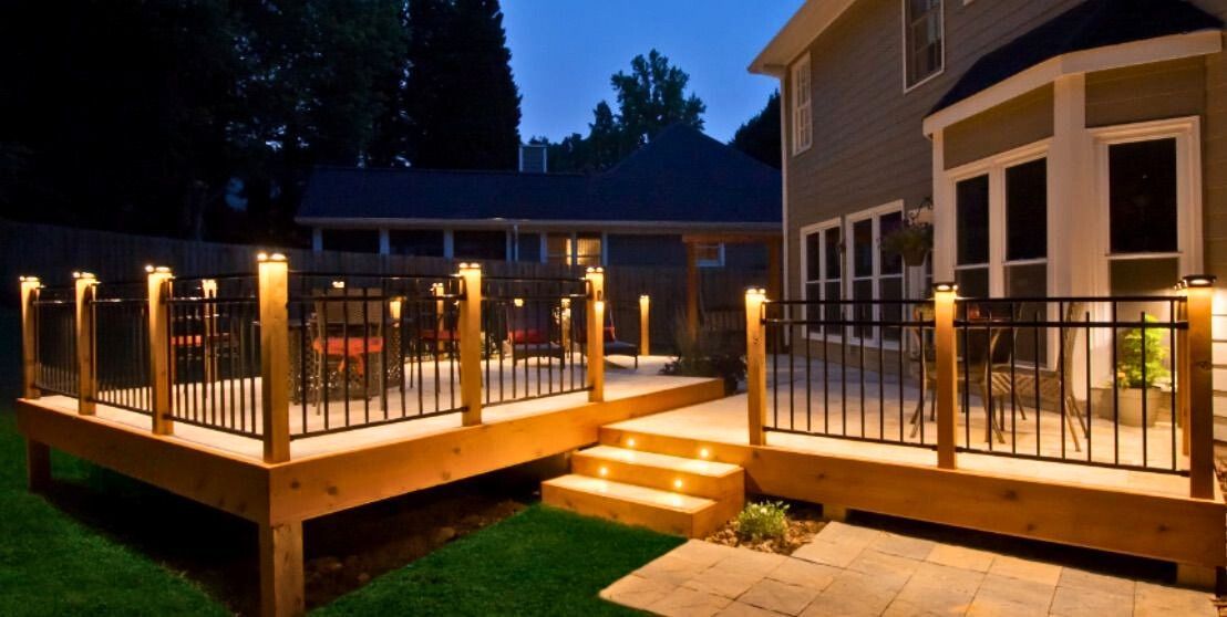 Deck illuminated with lights at dusk. The wooden deck has black railings, steps and backyard of a house.