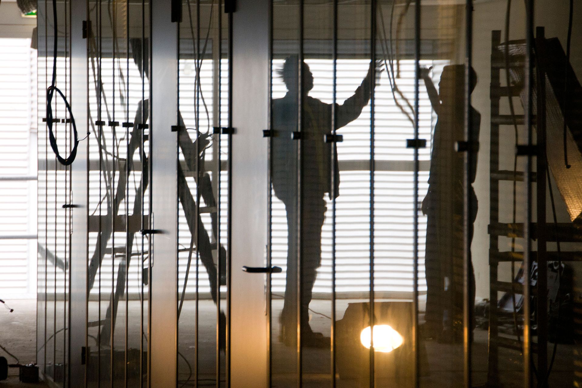 Silhouettes of two people on a ladder, adjusting wires near a window with blinds.
