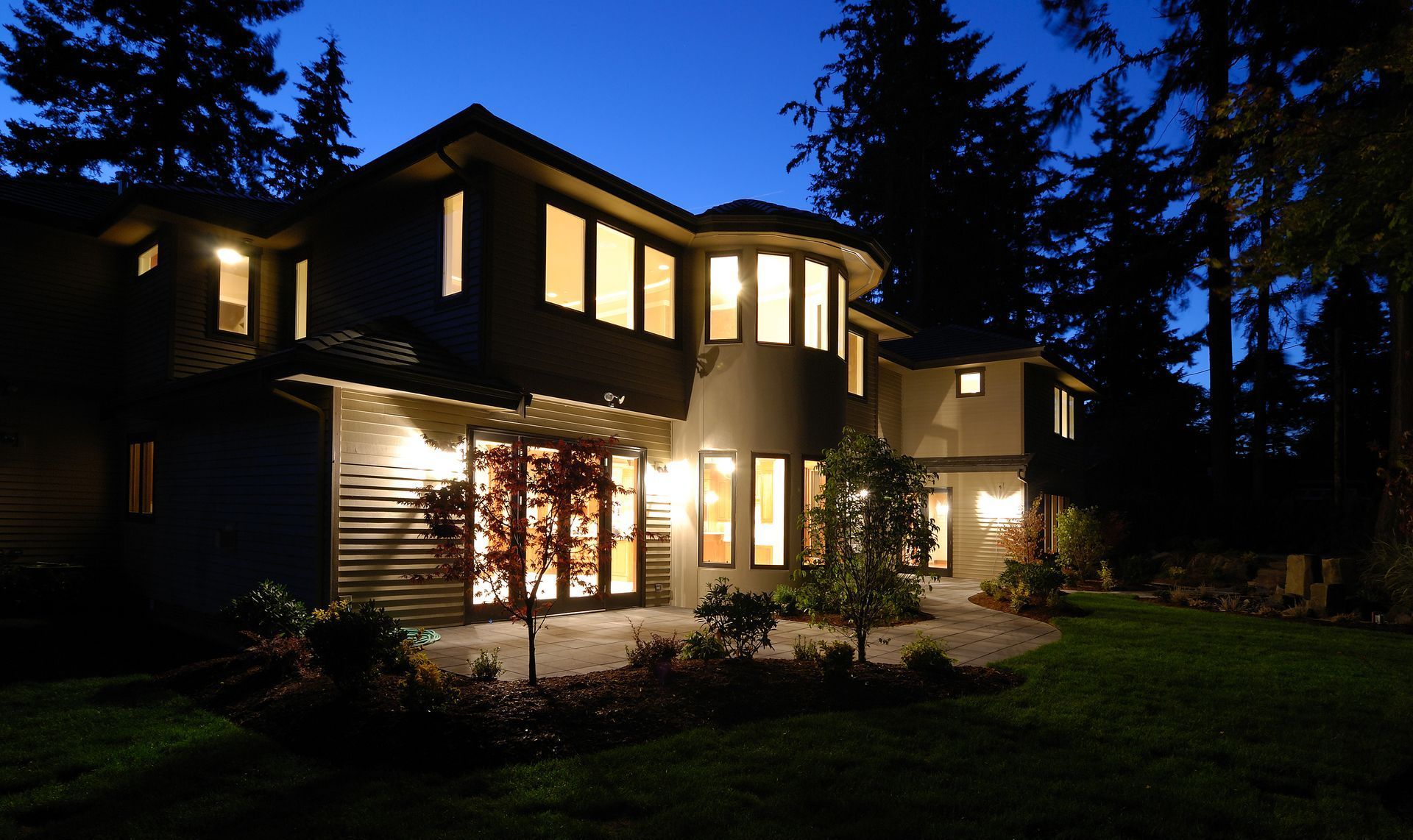 Two-story house at dusk, lit interior lights, surrounded by trees and a grassy yard.