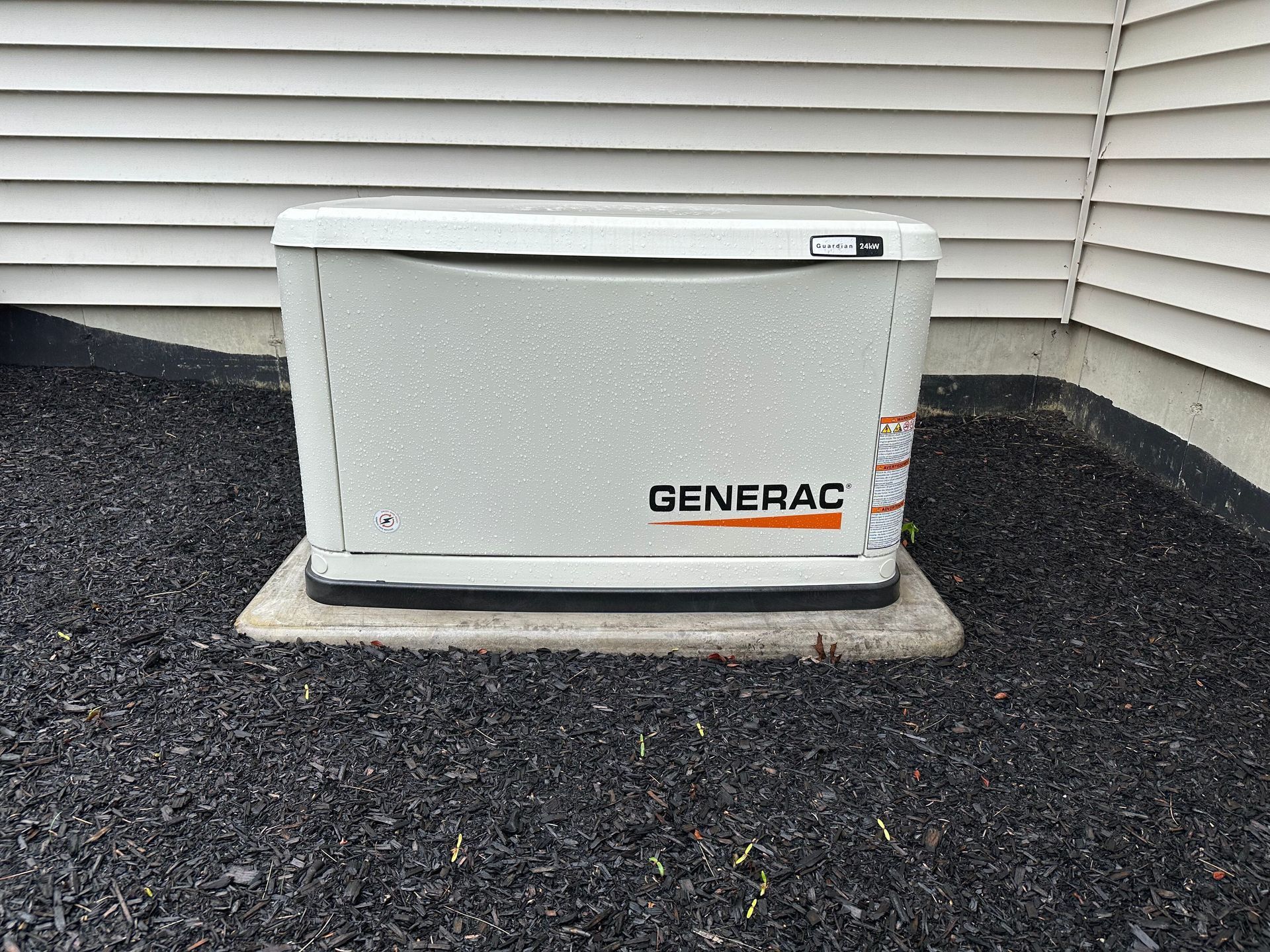 A Generac home standby generator on a concrete pad, surrounded by black mulch, beside a house with white siding.