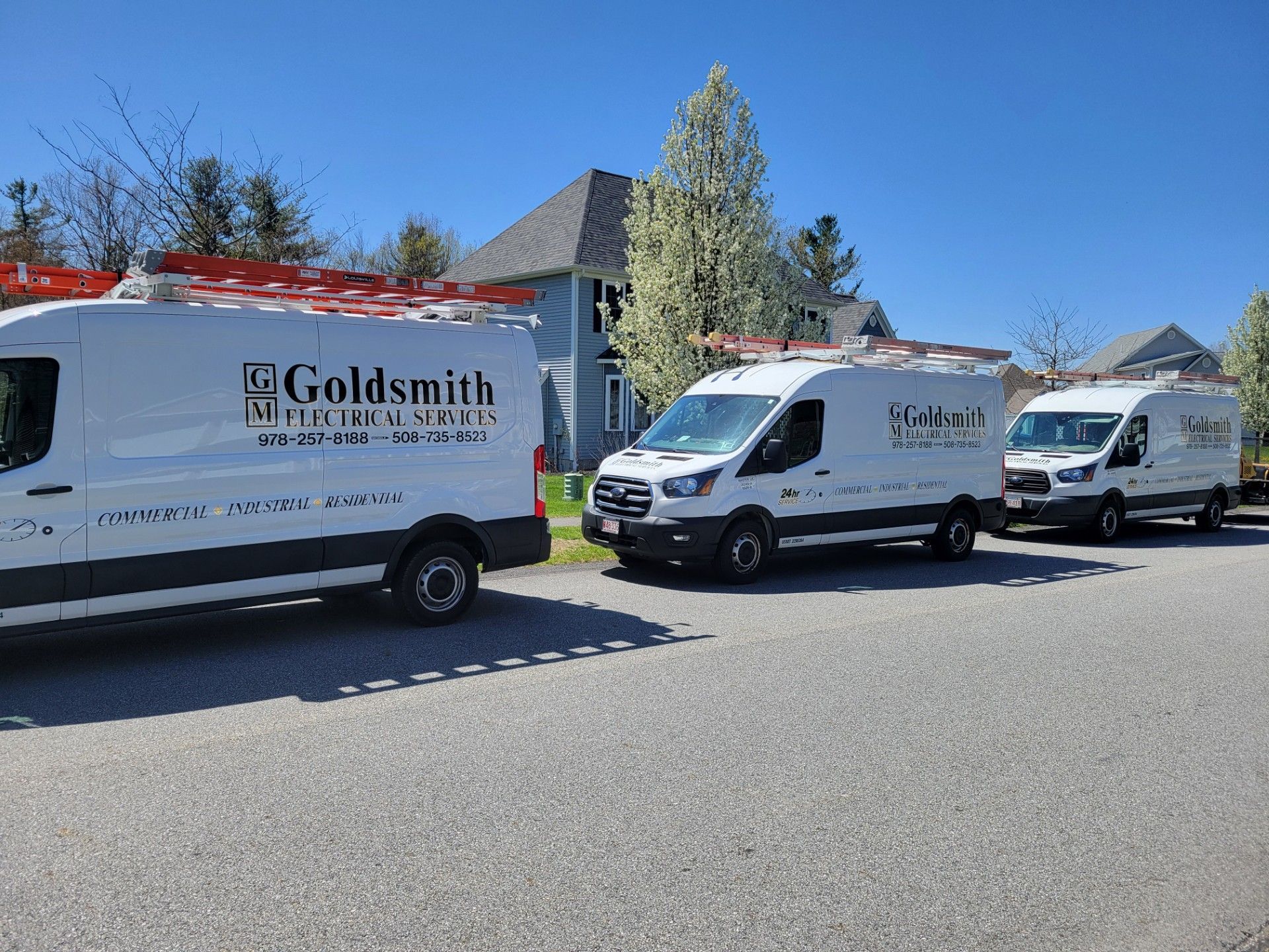 Three white Goldsmith Electrical Services vans parked on a street in front of a blue house, sunny day.
