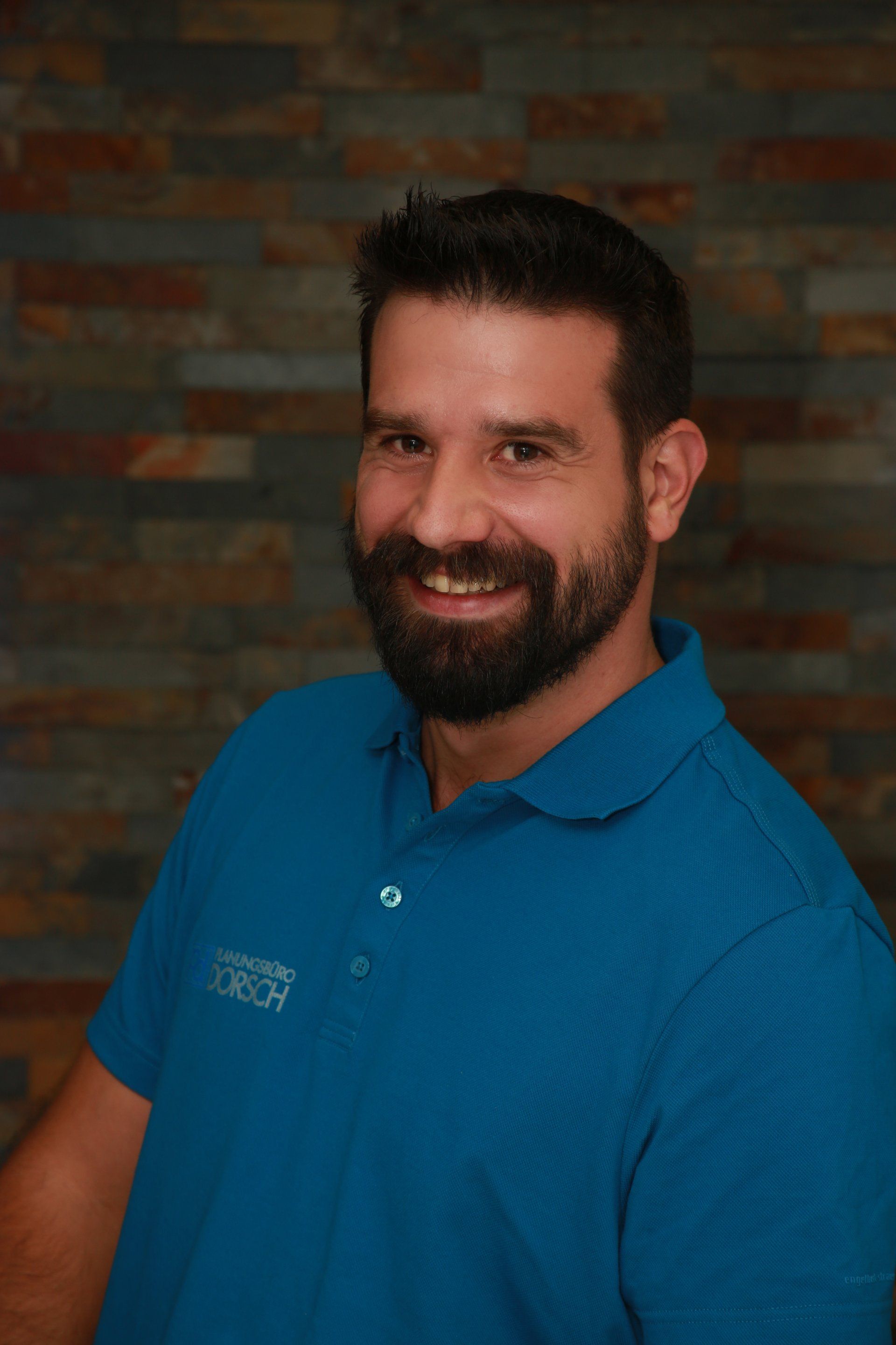 Ein lächelnder Mann in einem blauen Poloshirt posiert vor einer Backsteinmauer.
