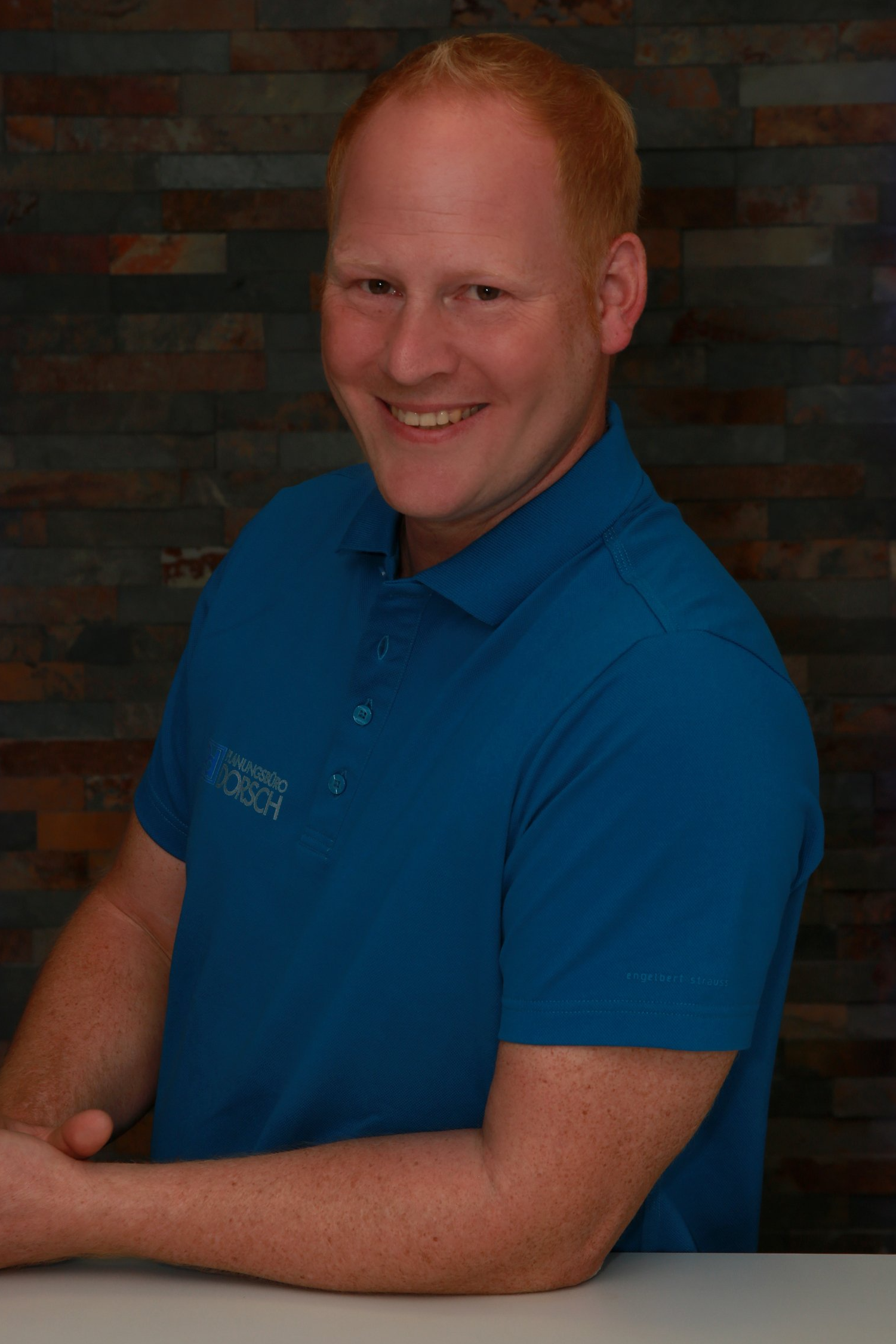 Eine lächelnde Person in einem blauen Poloshirt posiert vor einer dunklen Backsteinmauer.