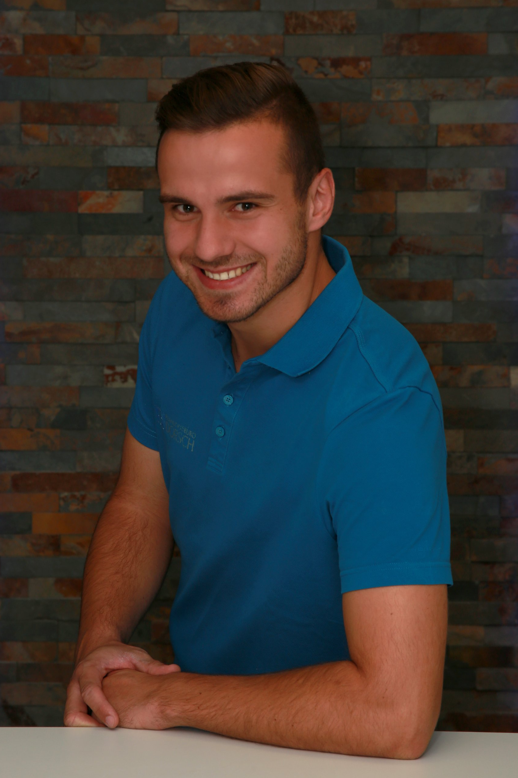 Ein Mann in einem blauen Poloshirt lächelt und lehnt an einem Tisch vor einer Backsteinmauer.