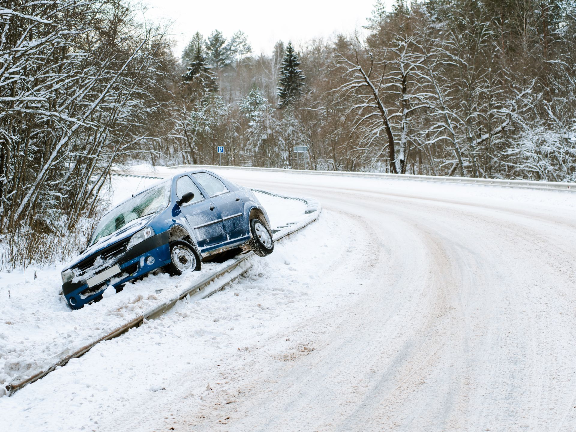 Blue car stuck in snow-filled ditch alongside a snowy, curving road.
