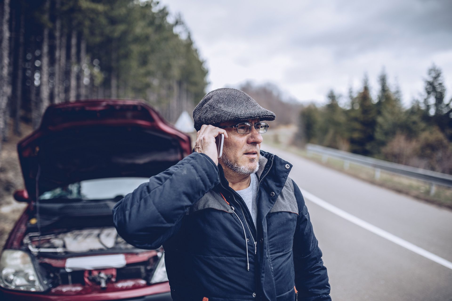 Man in a flat cap standing by a broken-down car with hood open on a roadside, looking ahead.