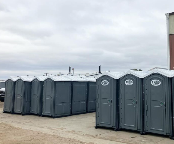 Rows of gray portable toilets lined up on a concrete surface against a cloudy sky.