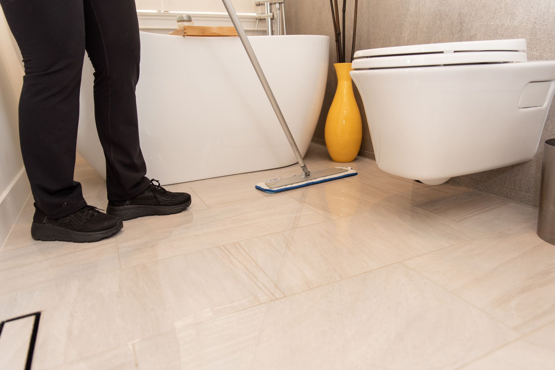 Person standing in a bathroom beside a mop, toilet, and yellow vase on a tiled floor
