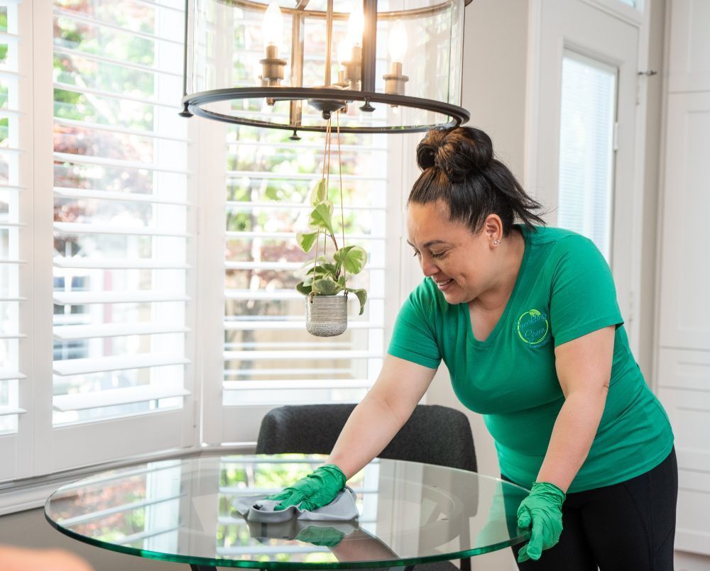 Sparking green technician in gloves cleaning a glass table in a bright room with window shutters.