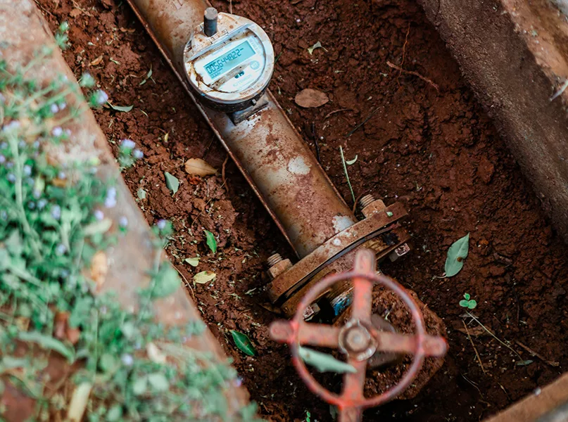 Rusty water meter and valve in a dirt-filled pit. The meter has a digital display and the valve is red.