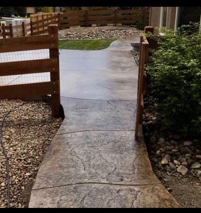 Stone walkway with a central strip leading through a fenced garden path, bordered by gravel and shrubs